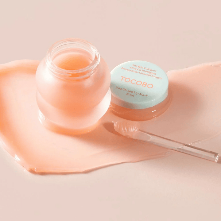 Prikazana je posoda z "Vita Glazed Lip Mask" iz Tocobo. Posoda je prozorna s svetlo turkiznim pokrovom in vsebuje sijočo, roza masko za ustnice. Majhna lopatica leži poleg posode, del maske pa je razporejen po podlagi. Izdelek je zasnovan za vlaženje in nego ustnic.