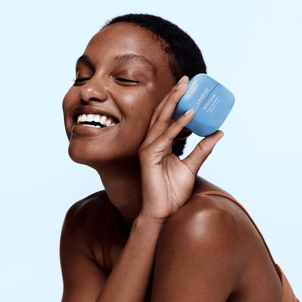 Nasmejana ženska drži moder lonček LANEIGE Water Bank Blue Hyaluronic Cream ob licu, s svetlo modrim ozadjem.