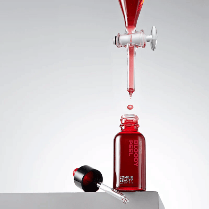 Steklenica Zombie Beauty Bloody Peel iz rdečega stekla, s pipeto poleg. Kapljica rdeče tekočine pada iz pipete zgoraj in je na poti v steklenico. Ozadje je svetlo sivo in poudarja dramatično barvo izdelka.