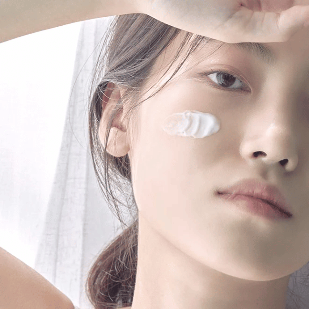 Bližnji posnetek mlade ženske s kančkom kreme na licu in dvignjeno roko nad čelom proti mehki podlagi.