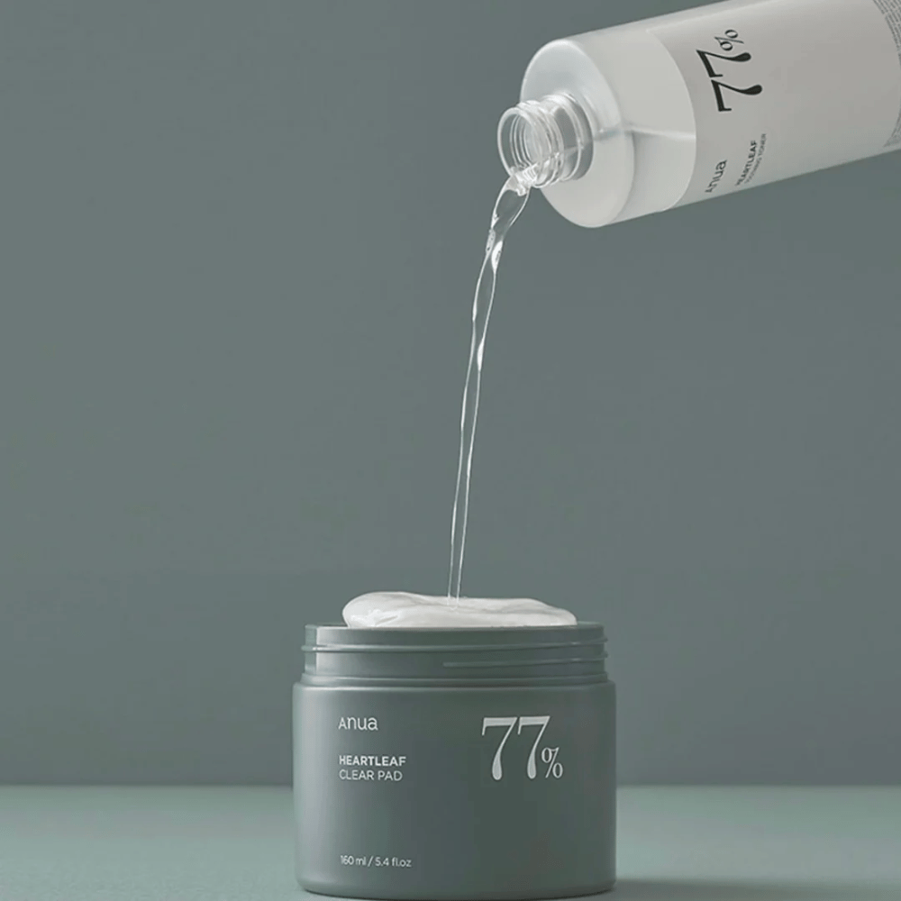 Prozorna steklenica naliva tekočino v siv kozarec z oznako "anua HEARTLEAF 77% Clear Pad", ki vsebuje kup belih blazinic za nego kože. Ozadje je preprosto in enobarvno v mehkem sivem tonu.