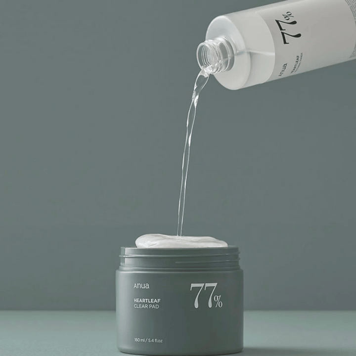 Prozorna steklenica naliva tekočino v siv kozarec z oznako "anua HEARTLEAF 77% Clear Pad", ki vsebuje kup belih blazinic za nego kože. Ozadje je preprosto in enobarvno v mehkem sivem tonu.