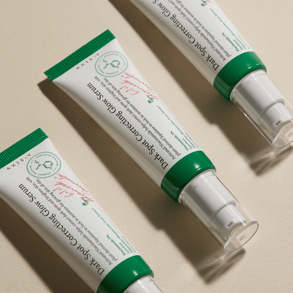 Tri tube Axis-Y Dark Spot Correcting Glow Serum z zelenim pokrovčkom in belo embalažo ležijo na svetli površini.