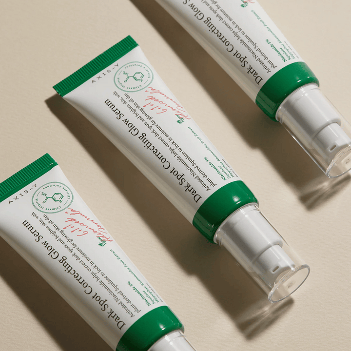 Tri tube Axis-Y Dark Spot Correcting Glow Serum z zelenim pokrovčkom in belo embalažo ležijo na svetli površini.