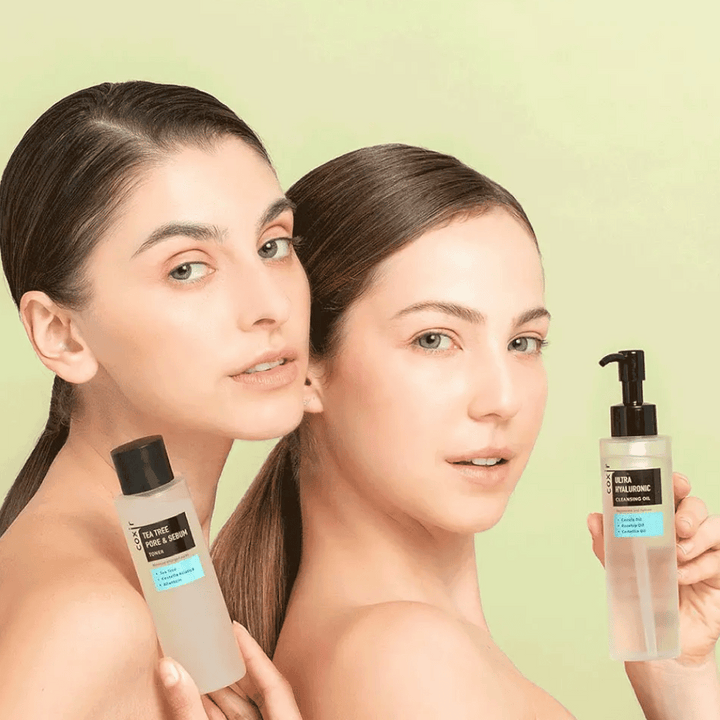 Dve ženski z golimi rameni pozirata s kozmetičnimi izdelki; ena drži steklenico z oznako "TEA TREE PORE & SEBUM", druga pa stekleničko s pumpico z oznako "ULTRA HYALURONIC CLEANSING OIL". Stojita pred svetlo zelenim ozadjem.