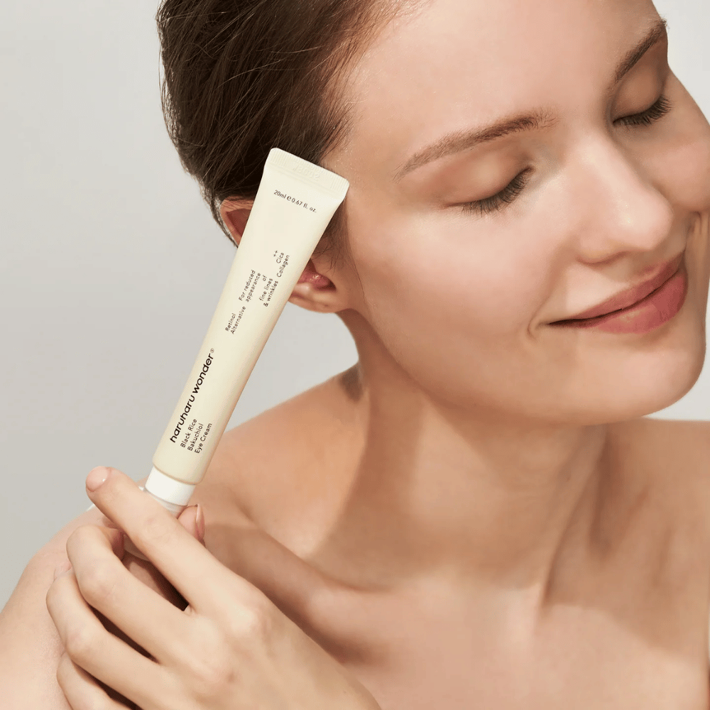 Nasmejana oseba z zaprtimi očmi drži tubo Haruharu Wonder Black Rice Bakuchiol Eye Cream proti sencu.