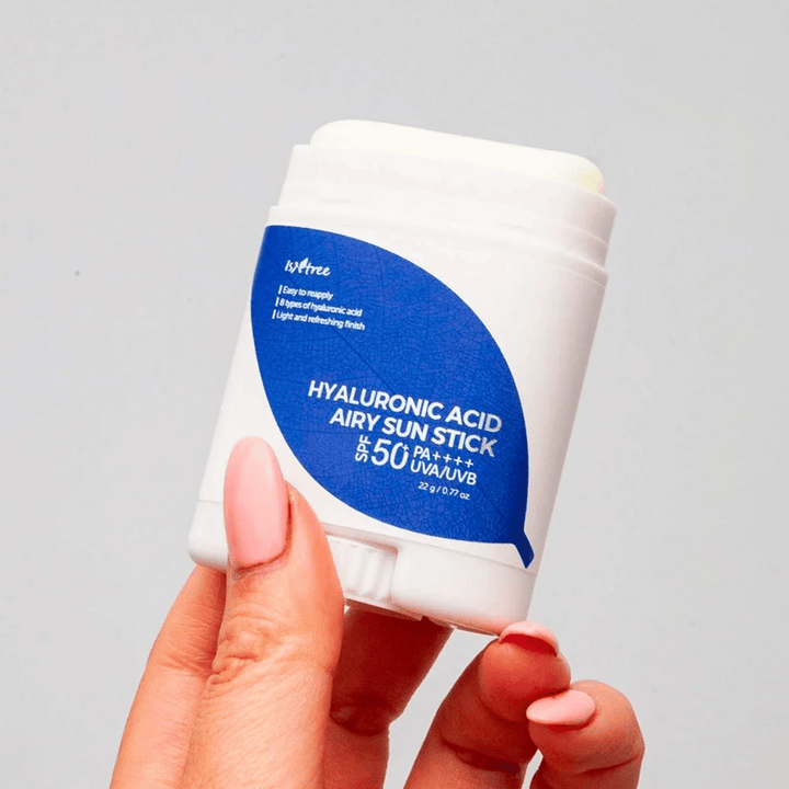 Roka drži belo sončno palico z modro etiketo blagovne znamke Isntree. Izdelek se imenuje "Hyaluronic Acid Airy Sun Stick" in ima zaščitni faktor SPF 50+ PA++++.