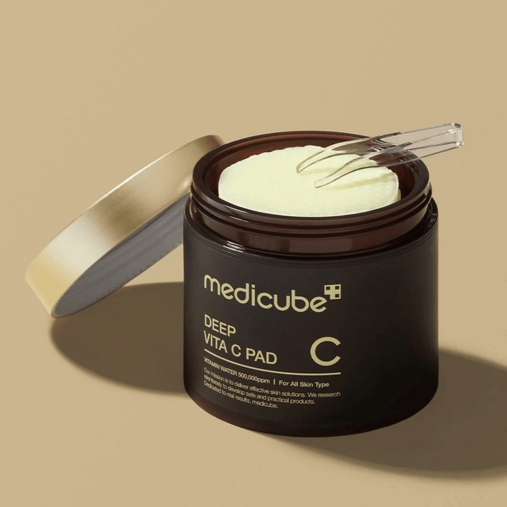 Kozarec z medicube Deep Vita C Pad. Kozarec je temno rjav z zlatim besedilom in belo, obrnjeno nalepko z informacijami o vitaminu C. Notranjost pokrova je zlata, prozorna pinceta pa leži na svetlo rumenih bombažnih blazinicah znotraj. Ozadje je enobarvno bež.