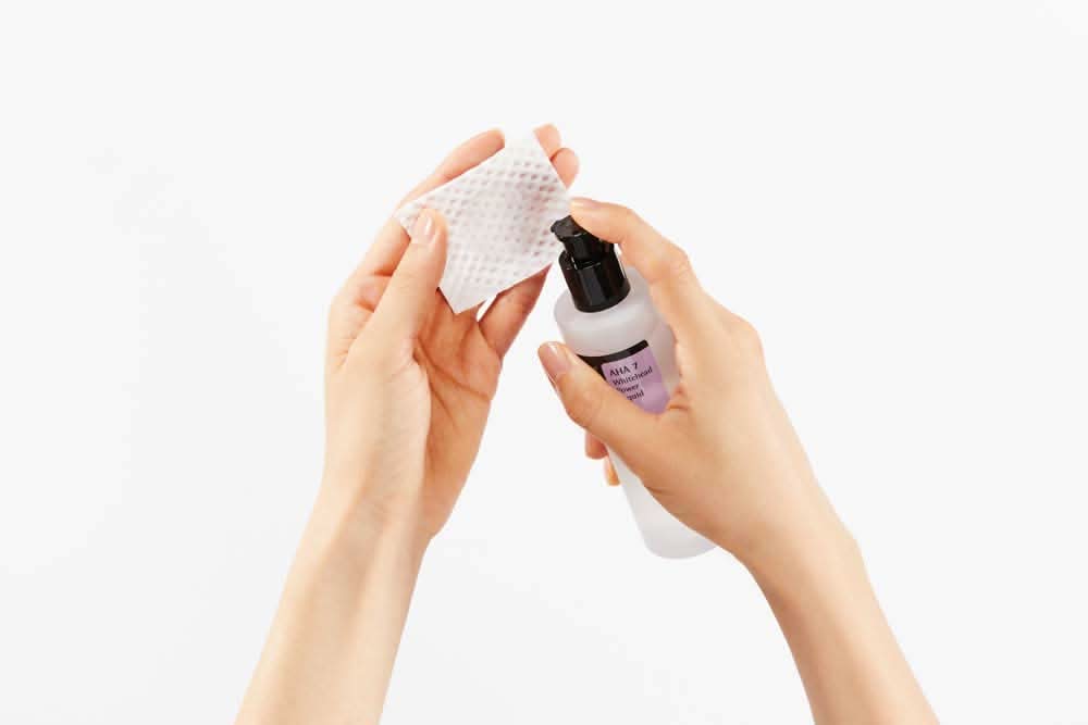 Roka, ki drži steklenico čistilnega sredstva s COSRX AHA 7 Whitehead Power Liquid 100ml za zdravljenje aken na beli podlagi.