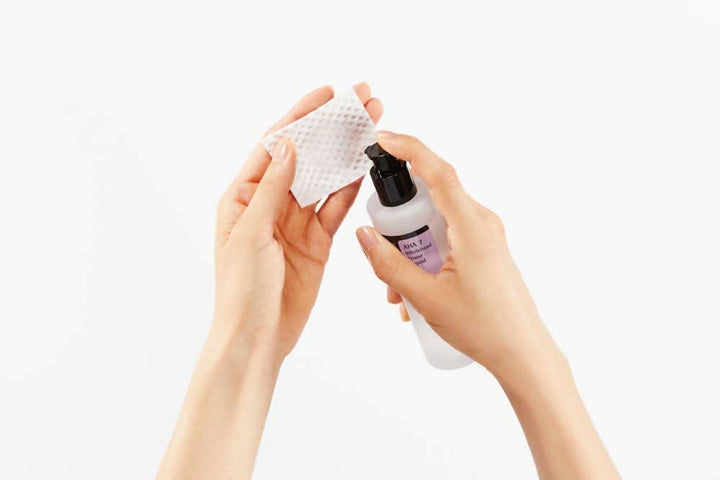 Roka, ki drži steklenico čistilnega sredstva s COSRX AHA 7 Whitehead Power Liquid 100ml za zdravljenje aken na beli podlagi.