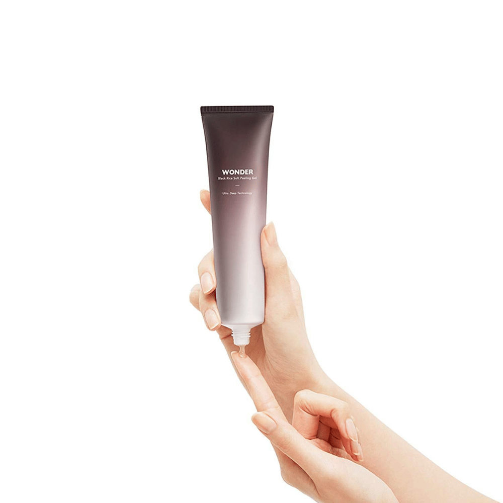 Ženska roka, ki drži tubo Haruharu Wonders Black Rice Soft Peeling Gel 100 ml na beli podlagi.