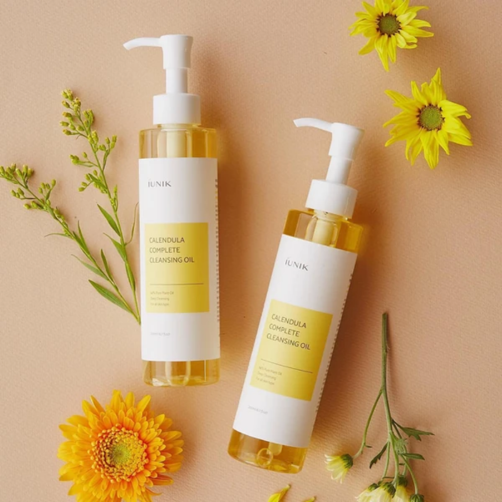 Dve steklenici iUNIK Calendula Complete Cleansing Oil 200ml z rumenimi cvetovi na bež ozadju, obogatena z rastlinskim oljem.