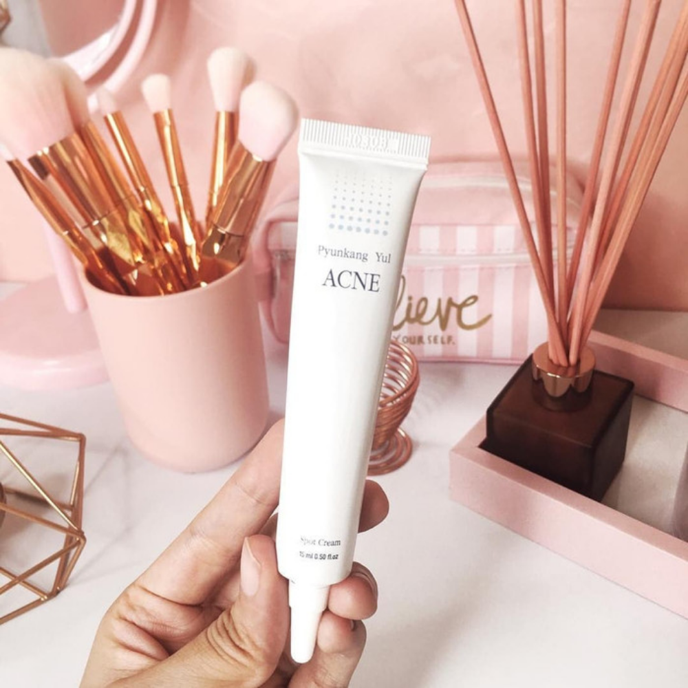 Oseba, ki drži tubo Pyunkang Yul Acne Spot Cream 15 ml pred rožnato mizo.