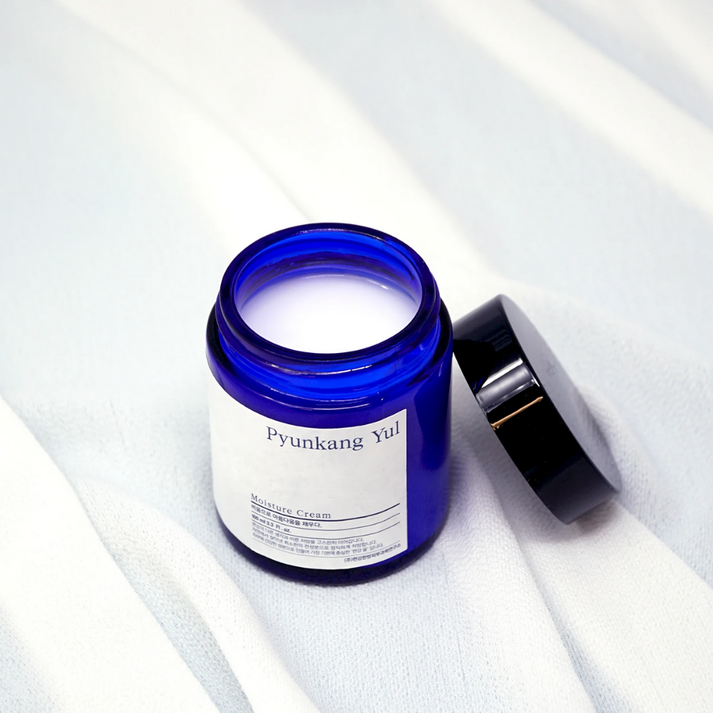 Moder kozarec s pokrovom, ki leži na beli krpi in prikazuje vlažilne lastnosti Pyunkang Yul's Moisture Cream 100ml.