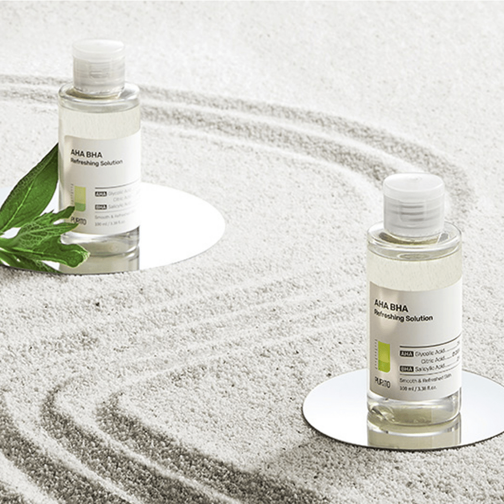 Dve steklenici PURITO AHA BHA Refreshing Solution 100 ml, ki stojita na peščeni površini.